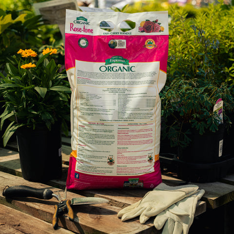Bag of Espoma Organic Rose-tone fertilizer on a wooden surface with gardening tools and plants in the background.
