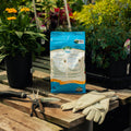 Fertilizer bag on a wooden table with gardening tools and gloves, surrounded by plants.