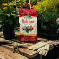 Bag of Espoma Organic Holly-tone fertilizer on a wooden table with gardening tools and gloves, surrounded by potted plants.