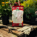 Bag of organic fertilizer on a wooden table with gardening tools and gloves, surrounded by plants.