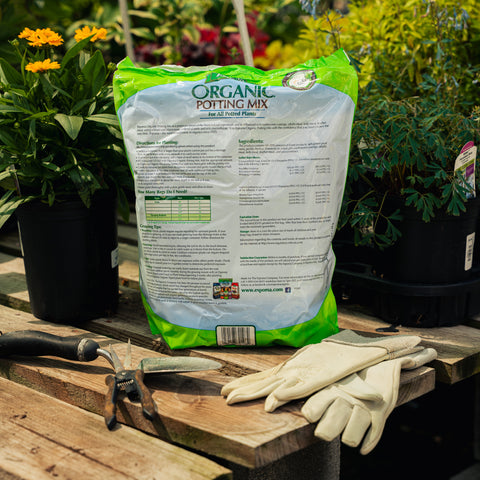 Bag of organic potting mix on a wooden table with gardening tools and gloves, surrounded by plants.