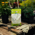 Bag of organic fertilizer on a wooden table with gardening tools and gloves, surrounded by potted plants.