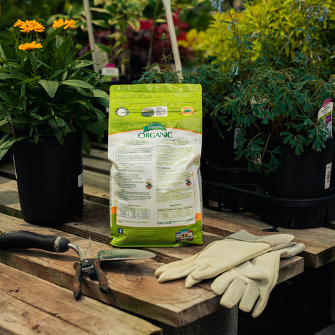 Bag of organic fertilizer on a wooden table with gardening tools and gloves, surrounded by potted plants.