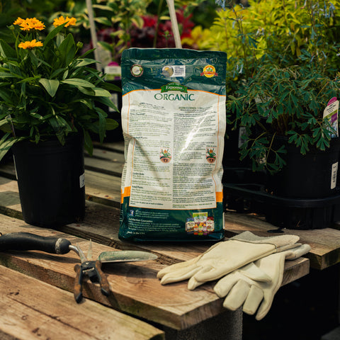 Bag of organic fertilizer on a wooden table with gardening gloves and tools, surrounded by plants.