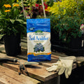 Bag of organic soil acidifier on a wooden table with gardening tools and gloves, surrounded by plants.