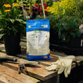 Bag of organic fertilizer on a wooden table with gardening tools and gloves, surrounded by plants.