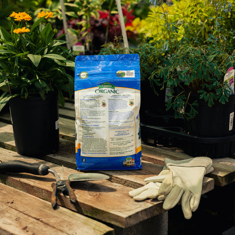 Bag of organic fertilizer on a wooden table with gardening tools and gloves, surrounded by plants.