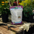 Bag of organic orchid mix on a wooden table with gardening tools and gloves, surrounded by plants.