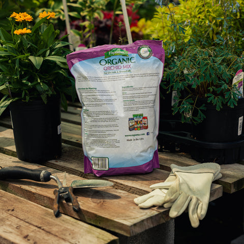 Bag of organic orchid mix on a wooden table with gardening tools and gloves, surrounded by plants.
