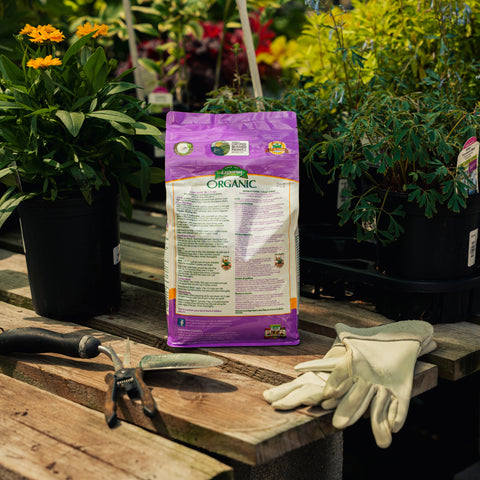 Bag of organic fertilizer on a wooden table with gardening tools and gloves, surrounded by plants.