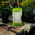 Bag of organic fertilizer on a wooden table with gardening tools and gloves, surrounded by plants.