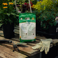 Bag of organic fertilizer on a wooden pallet with gardening tools and gloves, surrounded by plants.