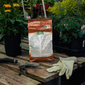 Bag of organic fertilizer on a wooden table with gardening tools and gloves, surrounded by plants.