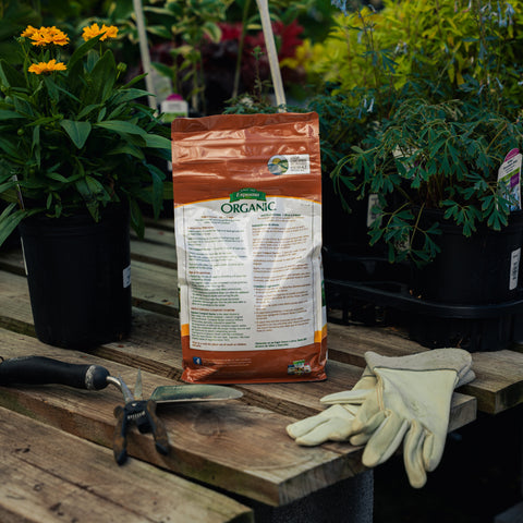 Bag of organic fertilizer on a wooden table with gardening tools and gloves, surrounded by plants.