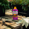 Bottle of 'Bloom!' fertilizer on a wooden surface with gardening tools and gloves in a garden setting.
