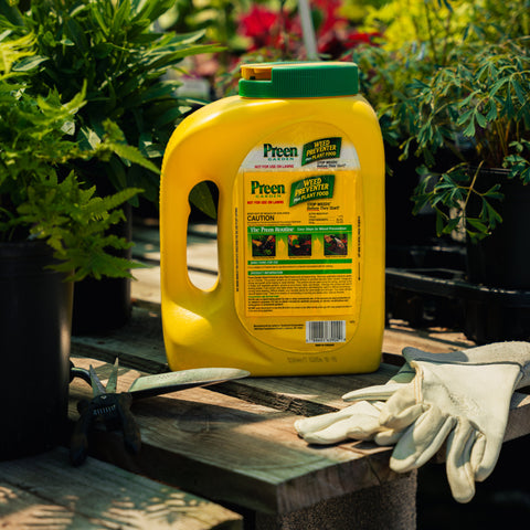 Yellow Preen weed killer container with gloves on a wooden surface surrounded by plants
