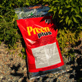 Red bag of Woodlace Preen Plus fertilizer on a gravel surface with green plants in the background