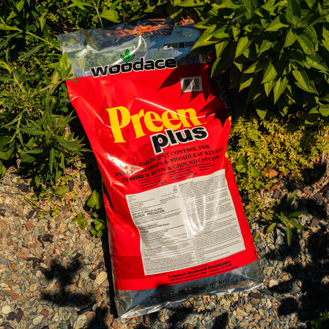 Red bag of Woodlace Preen Plus fertilizer on a gravel surface with green plants in the background