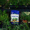 A bag of Garden Food fertilizer stands on a black nursery shelf surrounded by green potted plants.