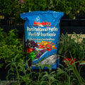 A blue bag of Lambert Horticultural Perlite standing upright in a garden setting surrounded by green plants.