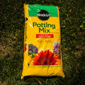 A bag of Miracle-Gro Potting Mix laid flat on green grass. The packaging is bright yellow with a green top banner, featuring the Miracle-Gro brand logo and colorful illustrations of flowers, including a large orange-red gerbera daisy.