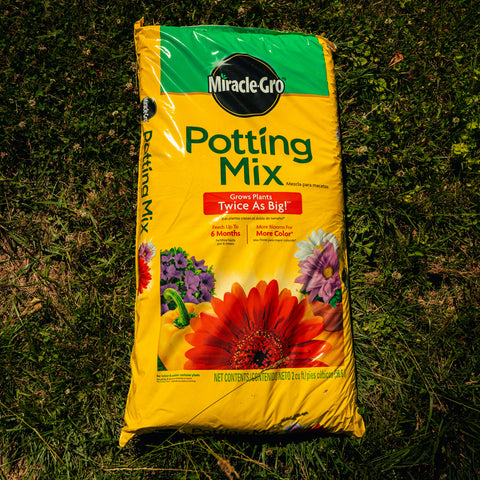 A bag of Miracle-Gro Potting Mix laid flat on green grass. The packaging is bright yellow with a green top banner, featuring the Miracle-Gro brand logo and colorful illustrations of flowers, including a large orange-red gerbera daisy.