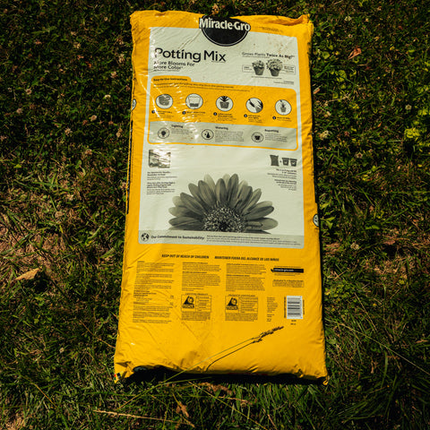 A large yellow bag of Miracle-Gro Potting Mix lies on green grass outdoors. The bag features a white label with a black daisy illustration and product details.