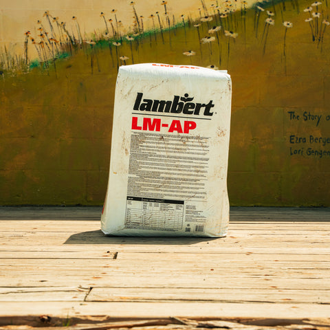 A white bag of Lambert LM-AP fertilizer stands on a wooden deck in front of a painted yellow wall with flowers.