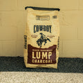 A 20 lb bag of Cowboy Brand Lump Charcoal, mixed hardwood flavor, standing upright against a beige painted brick wall on a dark asphalt surface.