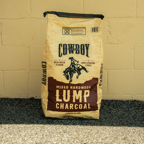 A 20 lb bag of Cowboy Brand Lump Charcoal, mixed hardwood flavor, standing upright against a beige painted brick wall on a dark asphalt surface.
