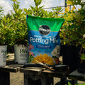 A bag of Miracle-Gro Moisture Control Potting Mix stands on a wooden table outdoors among potted plants. Gardening tools rest on the table, and other plant pots are visible in the background.