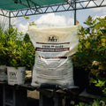 Bag of Twin Oaks Composted Manure in a garden setting with plants and a greenhouse.