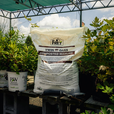 Bag of Twin Oaks Composted Manure in a garden setting with plants and a greenhouse.