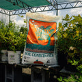 A large bag of Leafgro Soil Conditioner stands on a cinderblock table in a greenhouse or nursery setting, surrounded by potted green plants and garden tools.