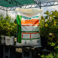 A large bag of Leafgro Soil Conditioner stands on a cinderblock table in a greenhouse or nursery setting, surrounded by potted green plants and garden tools.