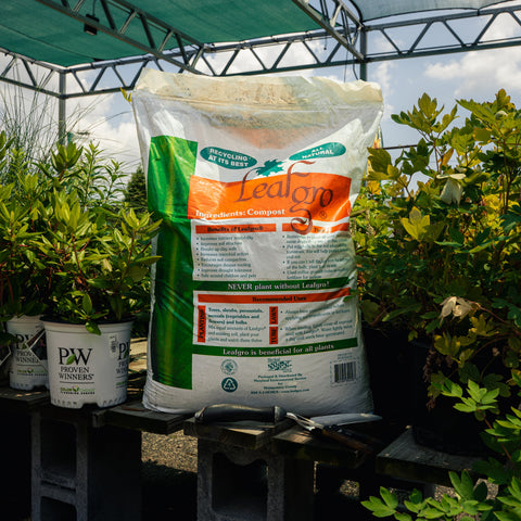 A large bag of Leafgro Soil Conditioner stands on a cinderblock table in a greenhouse or nursery setting, surrounded by potted green plants and garden tools.
