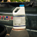 A white plastic jug of Cutrine Plus Aquatic herbicide with a blue cap, sitting on a stone pedestal in front of a black pond liner.