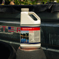 A white plastic jug of Weedtrine D aquatic herbicide with a red, white, and blue label, sitting on a black surface in front of a black tarp.