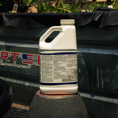 A white plastic jug of Weedtrine D aquatic herbicide with a red, white, and blue label, sitting on a black surface in front of a black tarp.