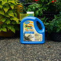 A blue plastic bottle of Roundup QuickFlow herbicide stands on a gravel surface between potted green plants.