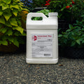 A white plastic jug of Cornerstone Plus liquid herbicide standing on a speckled stone surface outdoors, with green plants and flowers in the background.