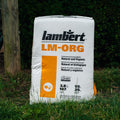 A large white bag of lambért LM-ORG Natural and Organic professional growing media stands on grass outdoors with trees in the background.