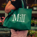 A woman carrying a green, mill-branded tote bag with a garden in the background