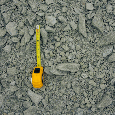 A yellow tape measure lies on a bed of gray gravel and small stones, showing its scale against the coarse material.
