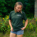 A woman wearing a dark green crewneck t-shirt with a white 'The Mill' logo on the chest, standing outdoors in a grassy field with yellow flowers and green foliage.