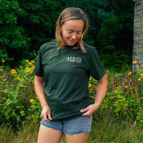 A woman wearing a dark green crewneck t-shirt with a white 'The Mill' logo on the chest, standing outdoors in a grassy field with yellow flowers and green foliage.