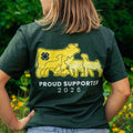 A woman shown from behind wearing a dark forest green t-shirt with a yellow graphic of livestock animals, with the text 'PROUD SUPPORTER 2026' printed below. The background is a blurred green garden with yellow flowers.