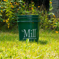 A dark green plastic bucket with a metal handle sits on a grassy lawn in front of a garden with yellow flowers. The bucket features the white printed text 'The Mill.'