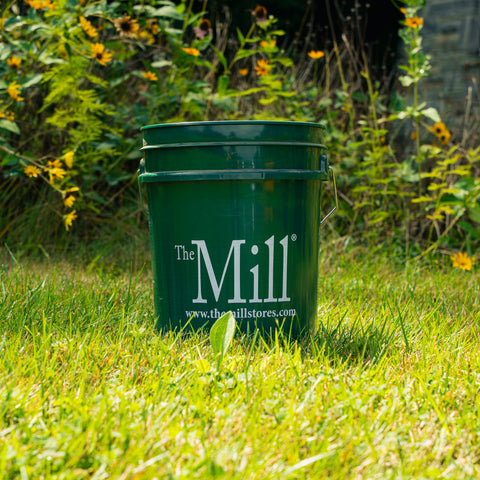 A dark green plastic bucket with a metal handle sits on a grassy lawn in front of a garden with yellow flowers. The bucket features the white printed text 'The Mill.'