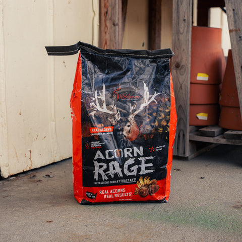 A bag of Acorn Rage Deer Attractant stands on a concrete floor in front of a garage door, with stacked terracotta pots visible in the background.